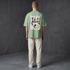 WOFF unisex sage green oversized terry tee with Stay Wild wolf graphic on the back, worn by a man in cream trousers.